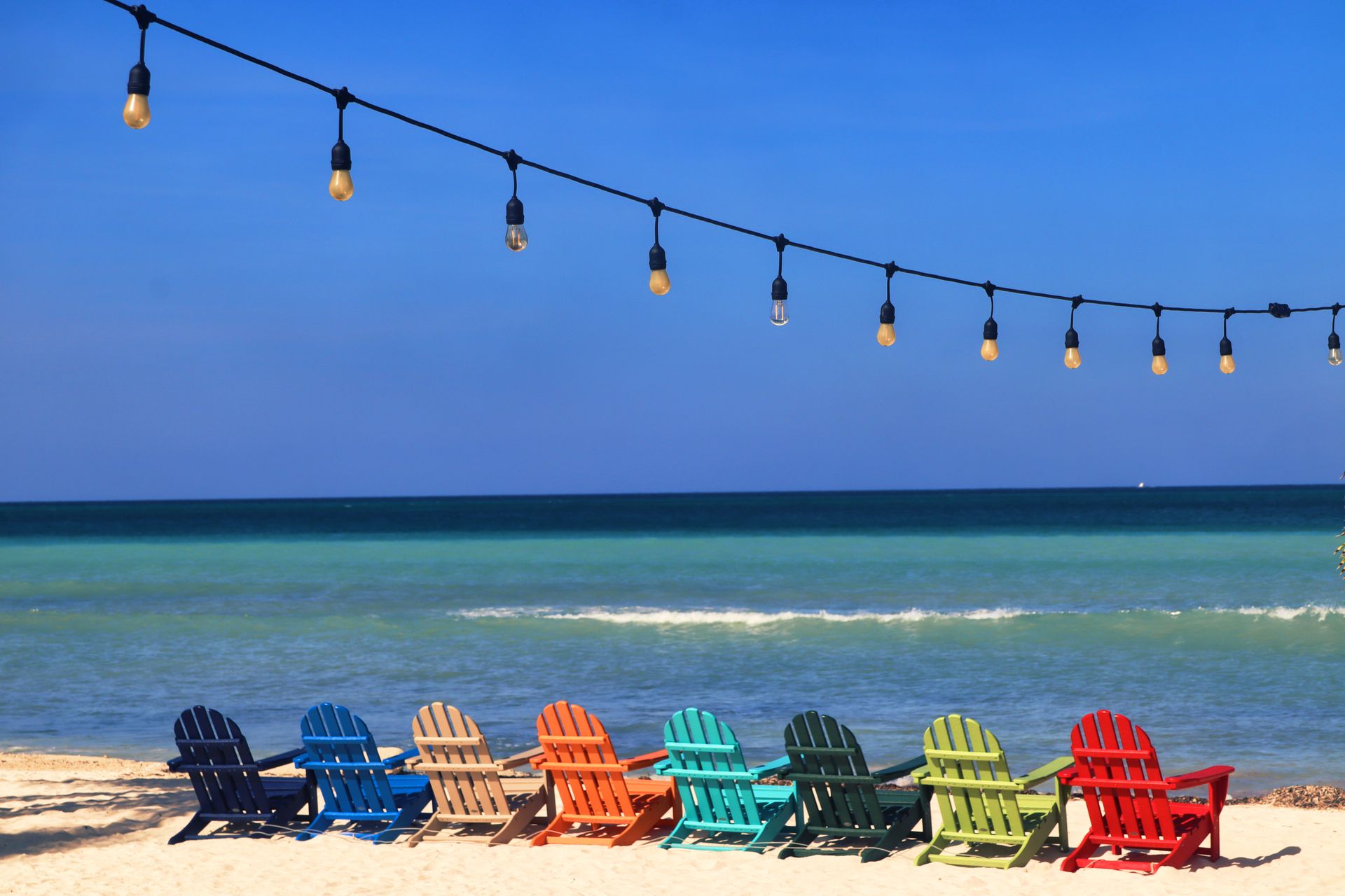 Colourful Adirondack chairs on a sandy beach beside turquoise Caribbean water in Aruba.