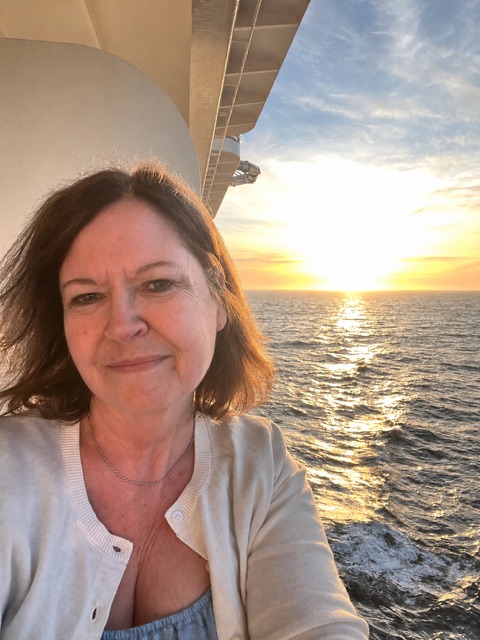 Pam Healy taking a selfie on a cruise ship balcony with the sun setting over the ocean