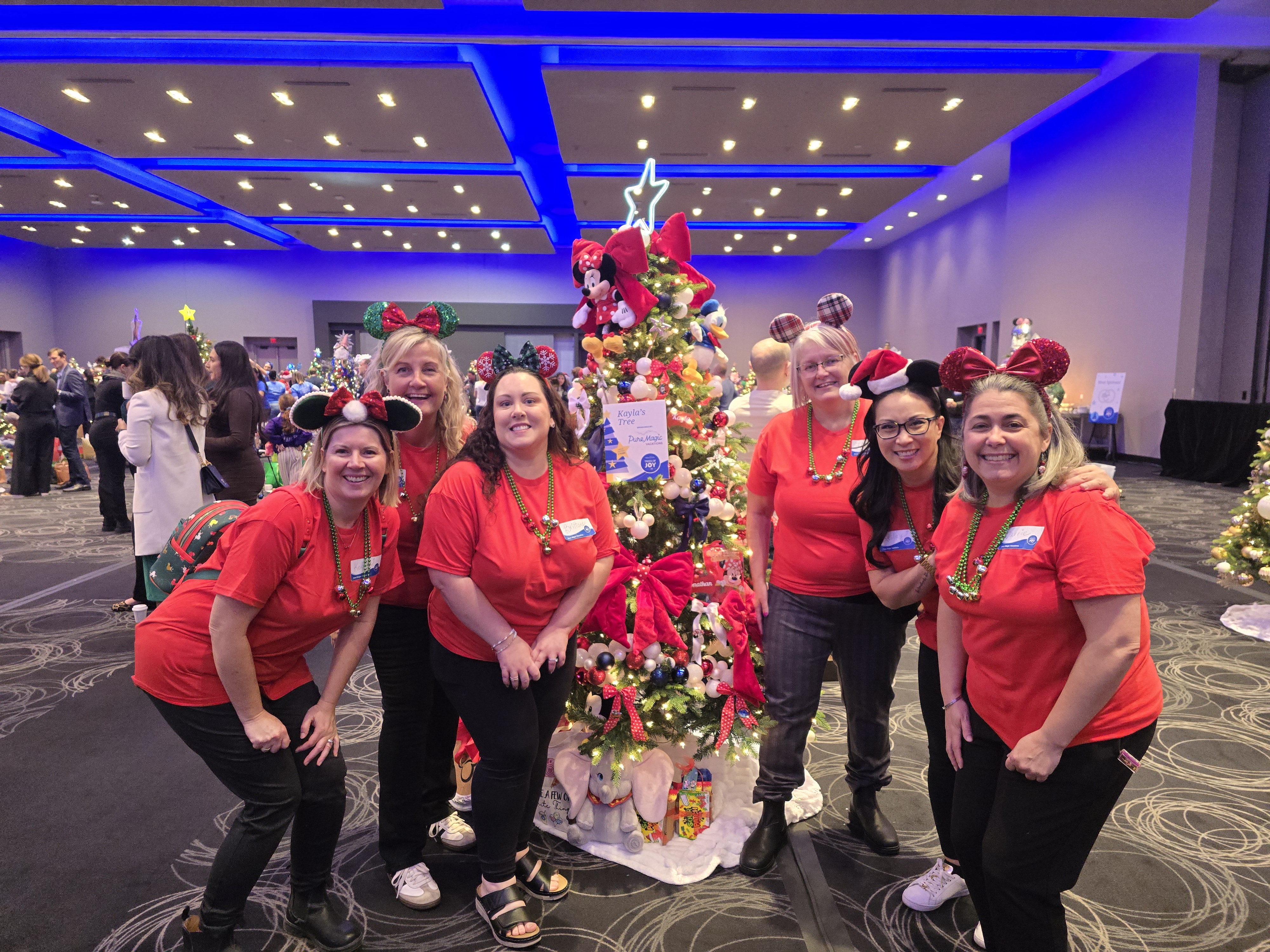 Six Pure Magic Vacations agents wearing red shirts pose beside a decorated Christmas tree at Trees of Joy.